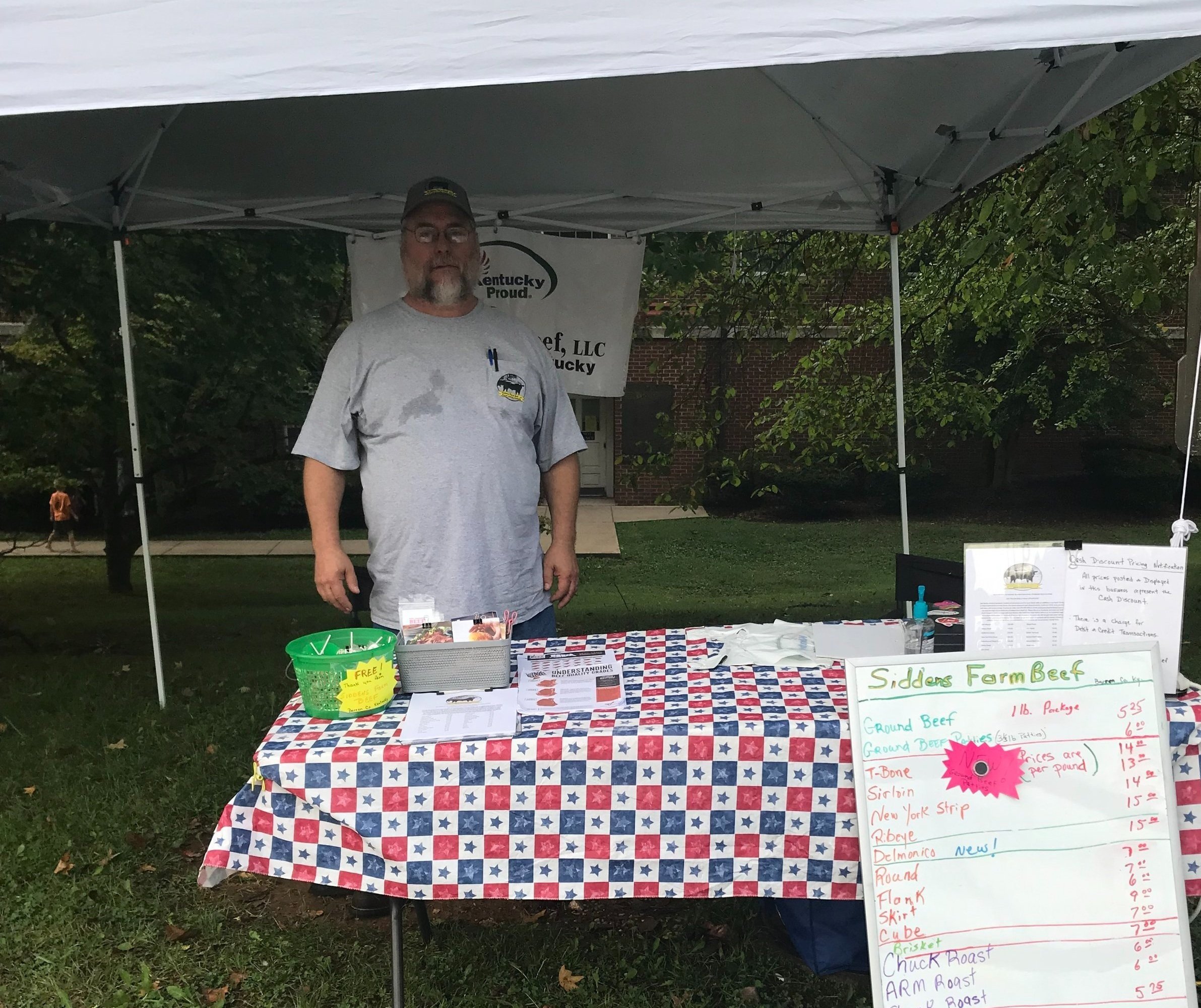 Robert+Siddens+-+IMG_1982 A vendor at a farmers market stands behind a table with a red, white, and blue tablecloth, showcasing various beef products from Siddens Farm Beef, LLC. The table features a price list for cuts of meat, including ground beef, T-bone, sirloin, and more. A green bucket labeled "FREE" is also visible, along with promotional materials and a Kentucky Proud sign in the background.