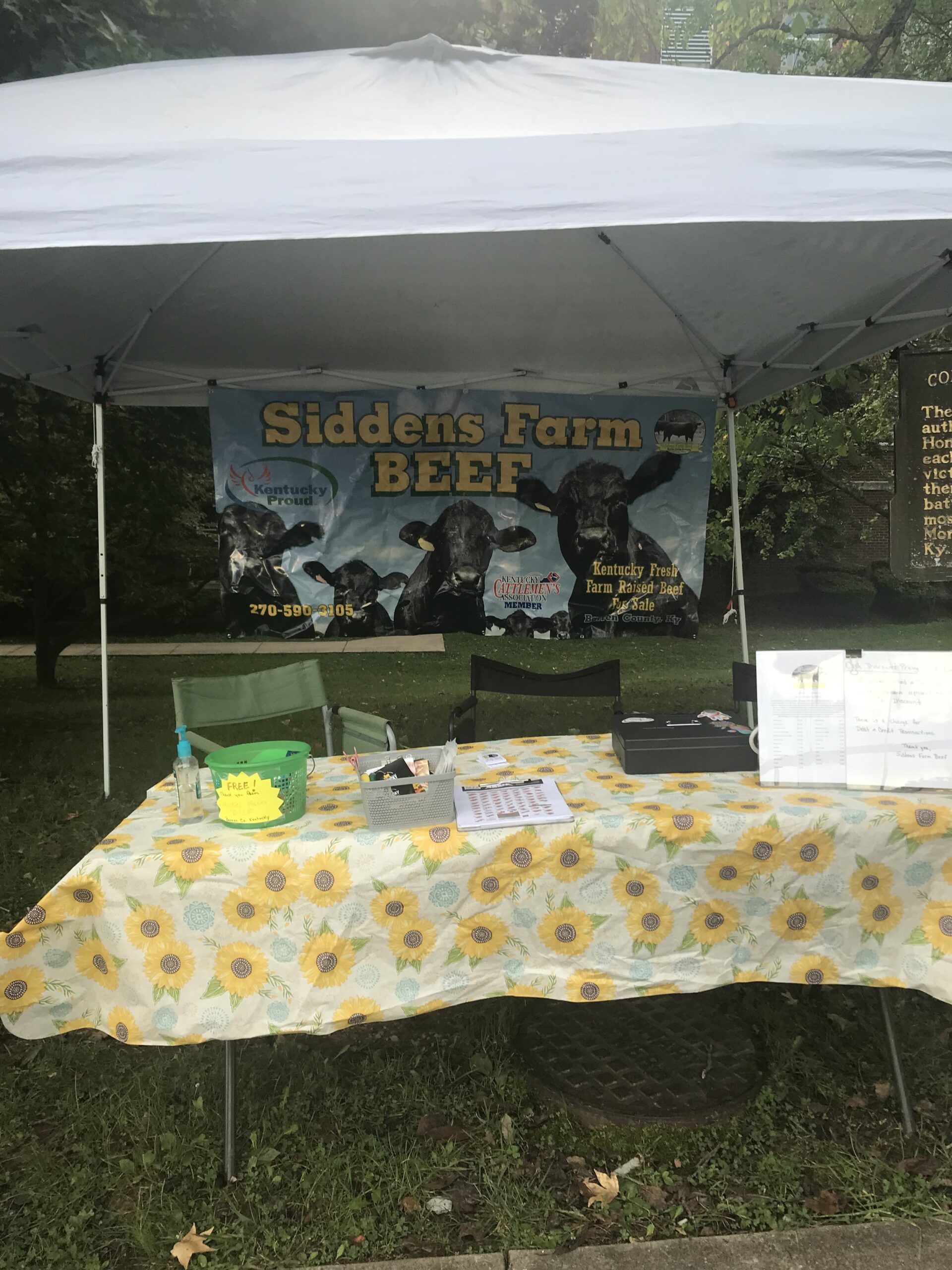 Robert+Siddens+-+IMG_2029 Tent setup for Siddens Farm BEEF featuring a colorful banner promoting Kentucky Proud farm-raised beef, with a table displaying promotional materials, a hand sanitizer, and a price list, all set in a grassy area.